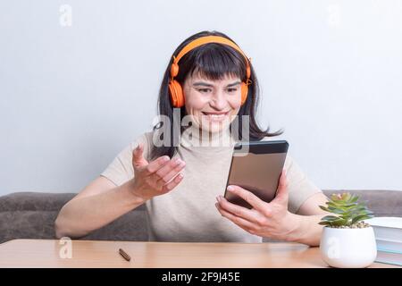 Felice sorridente allegra bella donna caucasica in cuffie arancioni che parlano online con gli amici nel tablet seduto al tavolo a casa. Donna co Foto Stock