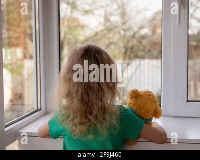 una bambina con il suo orsacchiotto guarda fuori la finestra al giardino. concetto di soggiorno a casa. fuoco selettivo Foto Stock