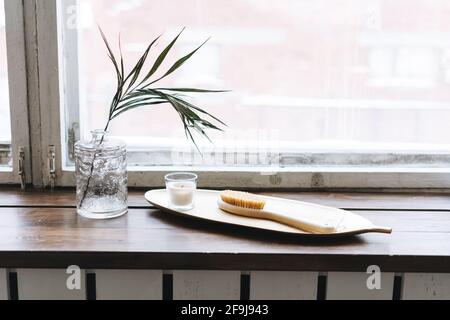 Dettagli interni, candela aromatica in vetro e spazzola in legno con setole naturali su vassoio in metallo su davanzale in legno Foto Stock