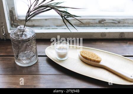 Dettagli interni, candela aromatica in vetro e spazzola in legno con setole naturali sul vassoio in metallo sul davanzale in legno Foto Stock