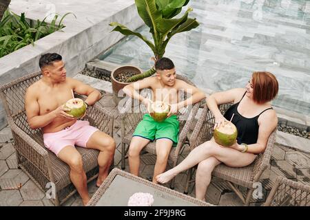 Una famiglia gioiosa che si siede su sedie di rattan dopo una nuotata in piscina, beve cocktail di cocco e discute di vacanza Foto Stock