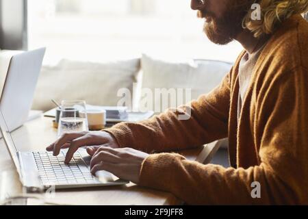 Minimo ritaglio di uomo bearded utilizzando il computer portatile mentre si lavora al bar tavolo illuminato dalla luce del sole in calde tonalità marrone, spazio di copia Foto Stock