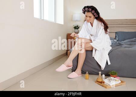 Giovane donna vietnamita in accappatoio che applica olio o siero idratante sulle gambe quando si siede sul letto in camera da letto Foto Stock