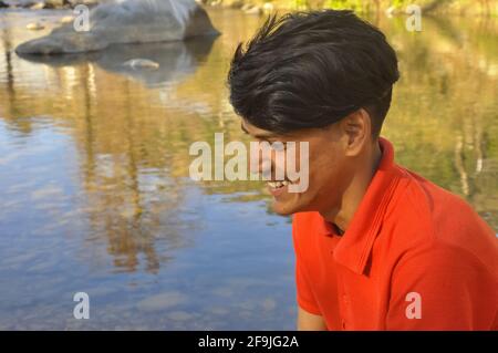 Primo piano della vista laterale di un giovane indiano sorridente con guardare lateralmente e seduto accanto al fiume, giovane uomo con tshirt di colore rosso, UN giovane adulto con la pelle imperfetta Foto Stock