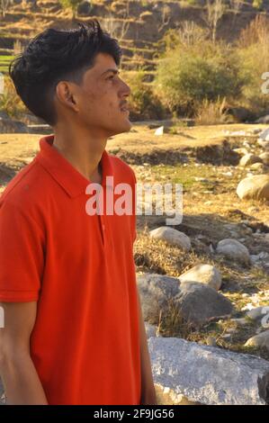 Ritratto di un giovane del sud asiatico in piedi all'aperto e guardando lateralmente, giovane uomo con tshirt di colore rosso, UN ragazzo adulto giovane con la pelle imperfetta Foto Stock