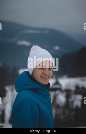 Ritratto candido di un ragazzo con un sorriso gioioso e infantile in un cappello bianco invernale e giacca blu. Ritratto reale di un adolescente sorridente naturale nel mon invernale Foto Stock