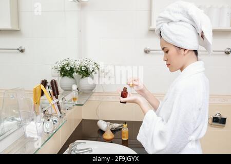 Bella giovane donna asiatica in accappatoio in piedi in bagno e. applicare siero idratante sulla sua faccia Foto Stock