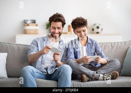 Intrattenimento e svago. Padre in competizione con il figlio in videogiochi online su smartphone, seduto insieme sul divano Foto Stock