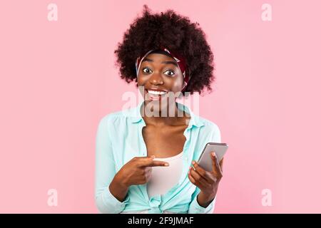 Donna africana entusiasta che punta il dito sullo smartphone su sfondo rosa Foto Stock
