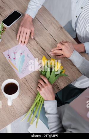 Vista dall'alto della donna seduta vicino a smartphone, biglietto di auguri e madre anziana con tulipani Foto Stock