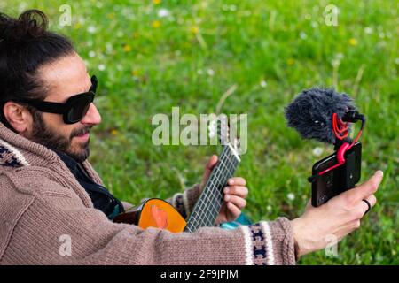 Hippy uomo con barba e occhiali che suonano la chitarra facendo un video se stessa all'aperto sull'erba Foto Stock