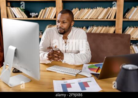 Freelance afro-americano concentrato seduto alla scrivania, utilizzando il computer, con una videoconferenza. Uomo d'affari serio ascoltare i colleghi a. Foto Stock