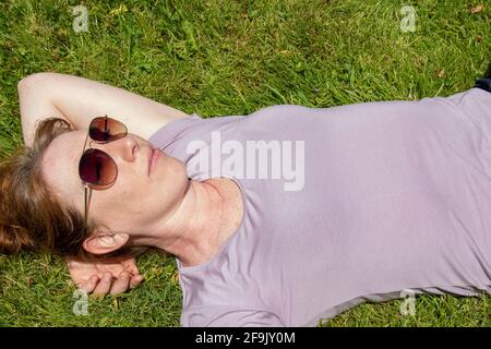 Donna di mezza età in occhiali da sole sdraiata sulla schiena godendosi il sole sulla sua faccia Foto Stock