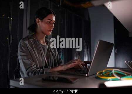 Ritratto di un tecnico di rete femminile che utilizza un computer portatile mentre si è seduti in una stanza server buia, copia spazio Foto Stock