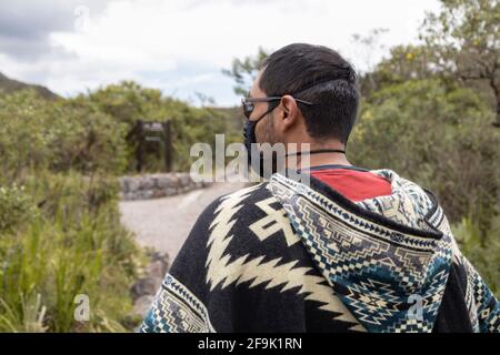 un uomo con un poncho, maschera e occhiali in piedi guardando un paesaggio con un percorso con alberi intorno ad esso, il turismo e lo stile di vita Foto Stock