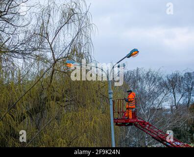 Dnepropetrovsk, Ucraina - 04.12.2021: Un operaio comunale in dispositivi di protezione che sostituisce le lampadine in una lampada da strada. Un lavoratore che ripara una lampada di strada di un bu Foto Stock