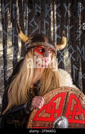 Una bella ragazza vichinga bionda in un casco cornuto è in piedi con uno scudo sullo sfondo del foresta e lo sfondo della scansione Foto Stock