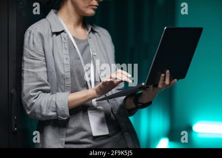 Ritratto troncato di un ingegnere di dati femminile che tiene un computer portatile mentre lavora con supercomputer nella stanza dei server, spazio di copia Foto Stock