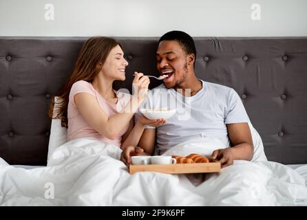 Felice coppia multirazziale che ha fatto colazione a letto, giovane donna che alimenta il marito con gustosi cereali Foto Stock