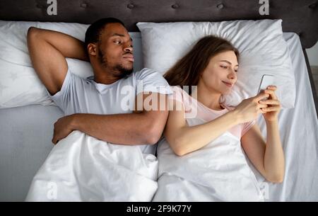 Irritato ragazzo nero che si sente geloso della sua ragazza bloccato in smartphone a letto, vista dall'alto. Concetto di phubing Foto Stock