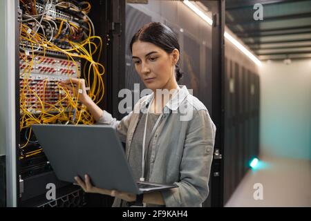 Ritratto di un tecnico di rete femminile che collega i cavi nell'armadietto del server mentre lavora con il supercomputer nel data center, spazio di copia Foto Stock