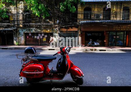 Uno scooter Vespa rosso parcheggiato si trova nella strada di fronte a una serie di vecchi edifici a Bangkok, Thailandia. Foto Stock