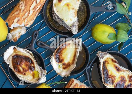 Ostriche cotte in casa in padelle di ghisa sul vecchio blu tavolo in legno decorato con limoni freschi con foglie di limone e. pane francese croccante Foto Stock