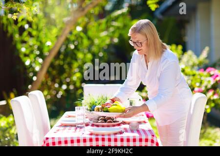 Donna che fissa tavolo all'aperto. Giardino estate divertimento. Barbecue in cortile soleggiato. Donna anziana a pranzo. Decorazione di festa. Femmina con frutta all'aperto. Foto Stock