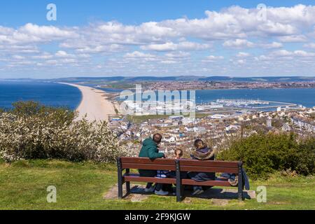 La famiglia in panchina gode di vedute della spiaggia di Chesil Bank e di Fortuneswell, Isola di Portland da Portland Heights, Portland, Weymouth Dorset, Regno Unito ad aprile Foto Stock