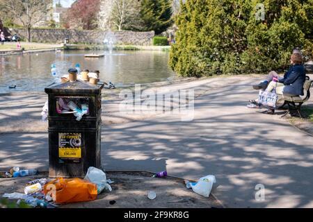 Pinner Regno Unito. Aprile 2021. Traboccante bidone dei rifiuti presso il ducklaghetto presso il Memorial Park, Pinner, Middlesex, Regno Unito. La coppia siede sulla panchina a lato. Foto Stock