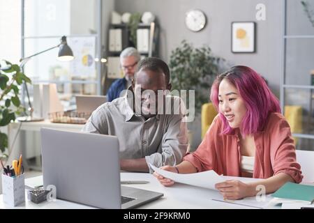 Giovane uomo d'affari africano seduto al tavolo con il suo asiatico colleghi e loro lavorano insieme al notebook Foto Stock