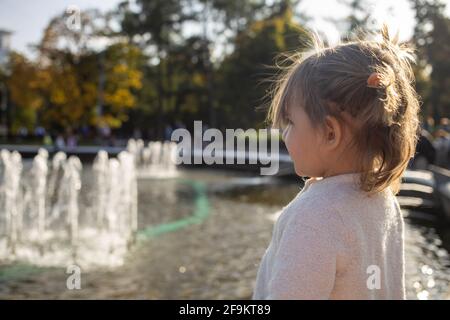 adorabile bambina guarda il laghetto con fontane nel parco in una giornata di sole. weekend a piedi in famiglia. passare il tempo con i bambini. attenzione artistica Foto Stock