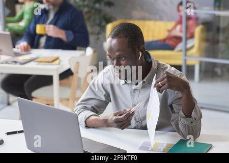 Giovane uomo d'affari africano che indica il documento nelle sue mani mentre avere una conferenza online su un computer portatile Foto Stock