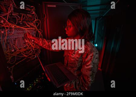 Vista laterale di una giovane donna afroamericana che collega i cavi mentre lavora con il supercomputer nella stanza del server illuminata da luce rossa, spazio di copia Foto Stock