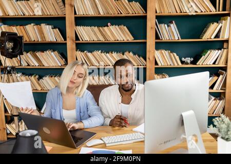 Colleghi di gara misti che utilizzano computer e computer portatile, facendo dei documenti, imparando grafici finanziari. Eleganti giovani colleghi che discutono di nuove idee per Foto Stock