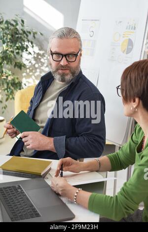 Uomo d'affari con portatore maturo in occhiali seduti al tavolo e. parlare con il dipendente in ufficio Foto Stock