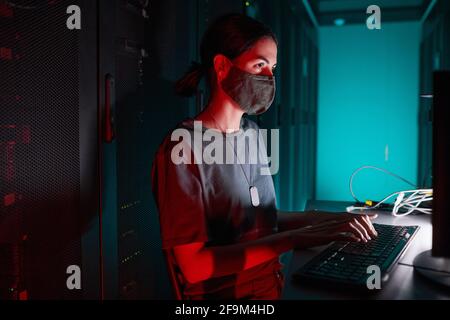 Ritratto di un tecnico IT femminile che indossa una maschera mentre si utilizza il computer e lavora in una sala server illuminata da luce rossa, spazio di copia Foto Stock