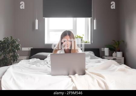 Bella ragazza caucasica sorridente si trova sul letto in camera da letto e utilizzare il computer portatile per il lavoro remoto. Concetti di comunicazione, tecnologia, lavoro a distanza in interni grigi Foto Stock
