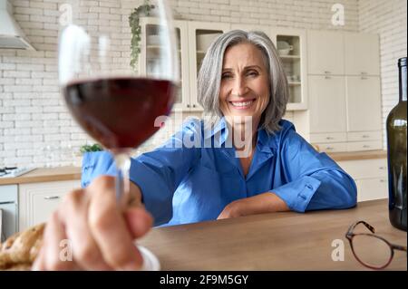 Media età 60 donna seduta in cucina bere vino online con la famiglia. Foto Stock
