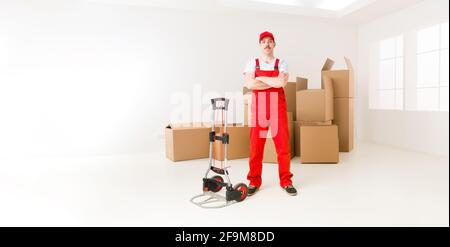 Giovani fiduciosi la consegna man standing nella nuova casa, tenendo le mani incrociate contro il suo petto, con le scatole di cartone in background Foto Stock