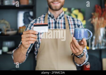 Uomo con bearded positivo che tiene in mano la tazza di ceramica e la carta di sconto mentre si posa al negozio di arredamento. Primo piano di mani di venditore caucasico in grembiule beige che mostra la carta di credito e la tazza blu alla macchina fotografica Foto Stock