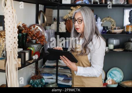 Affascinante donna anziana che usa un tablet digitale mentre fa l'inventario al negozio di arredamento. Donna di vendita competente con capelli grigi che indossa occhiali, camicia bianca e grembiule beige. Foto Stock