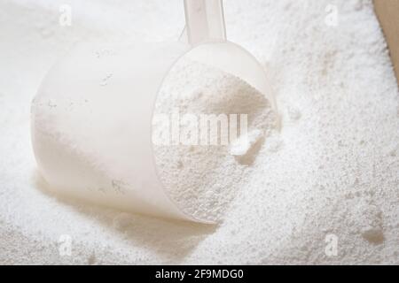 Primo piano della paletta in plastica trasparente utilizzata per misurare la polvere di detergente per bucato bianca in una scatola di cartone. Immagine luminosa con dettagli in polvere macro Foto Stock