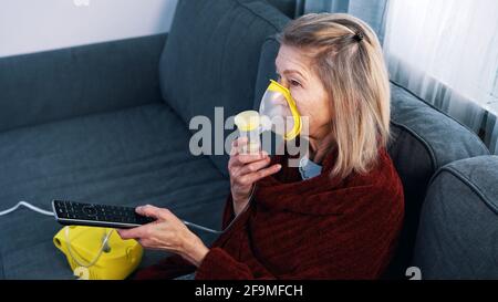 Donna anziana vulnerabile in quarantena che usa l'inalatore di ossigeno e cambia canel in tv. Foto di alta qualità Foto Stock