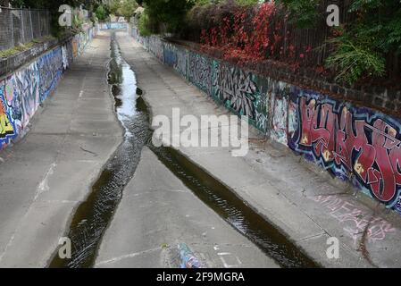 Un canale suburbano a Brighton Est in autunno, con acqua che lo percorre. Le pareti del canale sono coperte di graffiti, tra cui suggestivi murales. Foto Stock