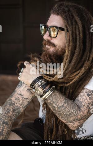 hipster ragazzo con dreadlock e tatuaggio seduto sulla spiaggia in thailan Foto Stock