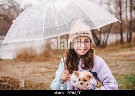 Bella ragazza con cane chihuahua sulla natura. Una ragazza adolescente e vestito cane in tempo piovoso sotto un ombrello. Cane cucciolo Foto Stock