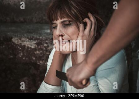 Primo piano della mano dell'uomo con un coltello, colpendo una donna spaventosa, di fronte al muro sporco. Ragazza che soffre di violenza, ha sangue di Th Foto Stock