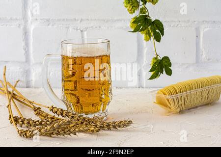 Tazza di vetro di birra con patatine, spighe di orzo e un ramo di luppolo su sfondo bianco. Foto Stock