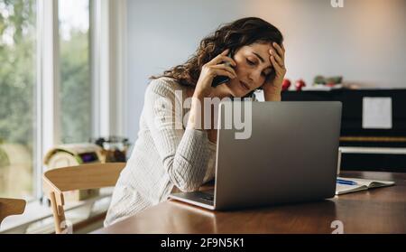 La donna si sente stanca mentre lavora da casa. Donna che guarda stressata seduta al tavolo con il computer portatile e che parla sul telefono cellulare. Foto Stock
